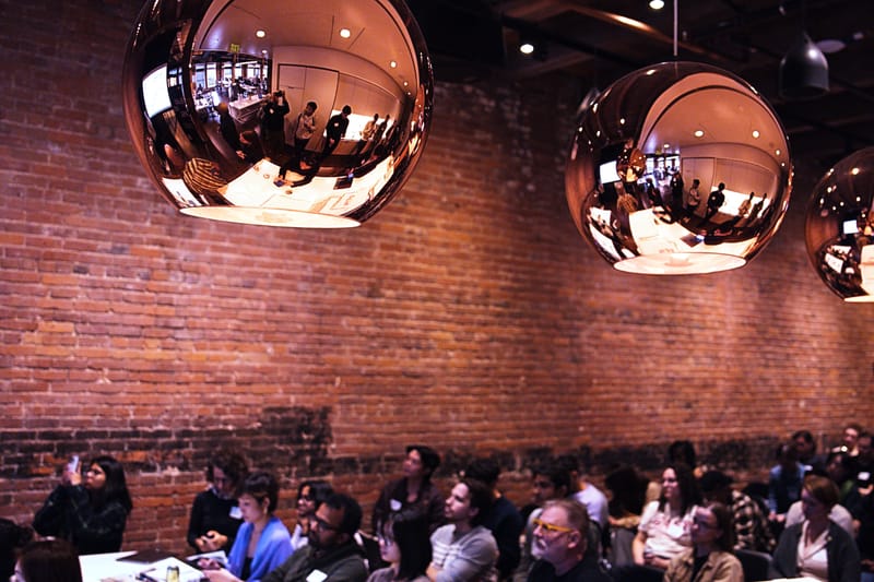 A full audience seated beneath large mirrored copper pendant lamps that reflect the room, against a long brick wall.