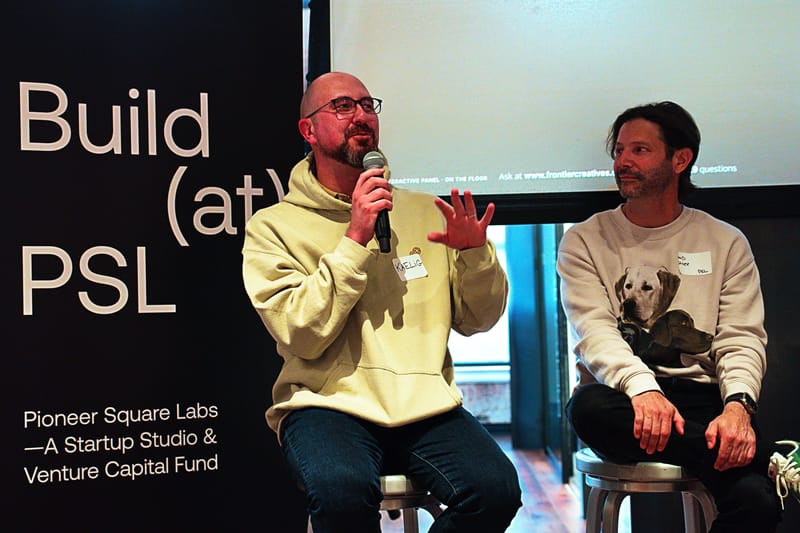 Two panelists on stools in front of the Build at PSL banner, one in a yellow hoodie speaking into a microphone.