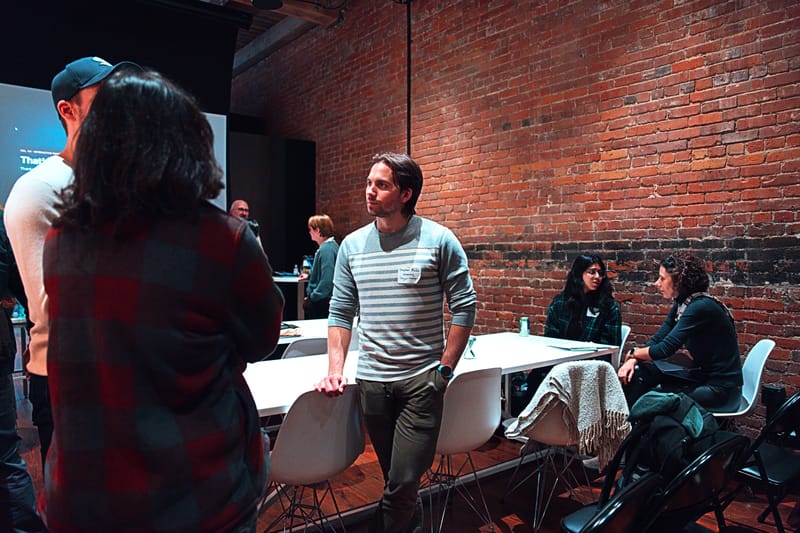 The emptying room after the talks, small groups still talking among the tables and chairs under the brick wall.